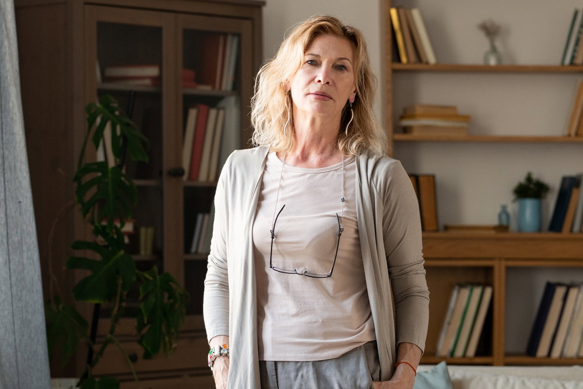 kobieta w wieku 50 plus blondynka w beżowym kardiganie stoi smutna w salonie