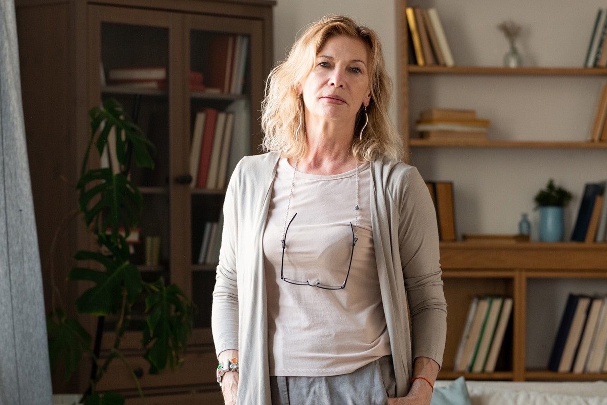 kobieta w wieku 50 plus blondynka w beżowym kardiganie stoi smutna w salonie