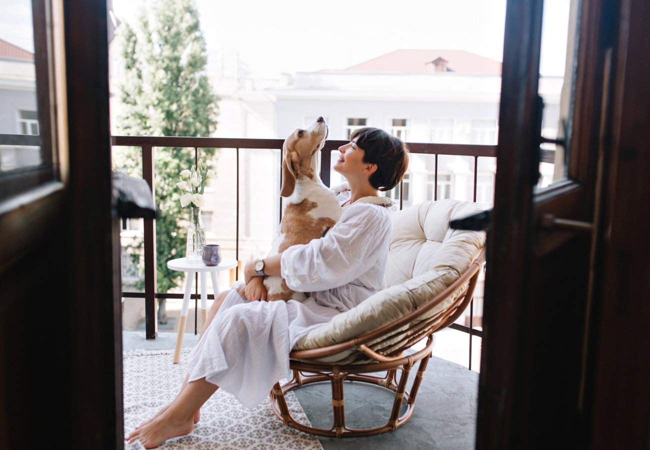 Relaxed young woman in white cozy bathrobe sitting with beagle dog on balcony in summer morning. Pretty brunette girl in wristwatch holding puppy on knees and smiling..