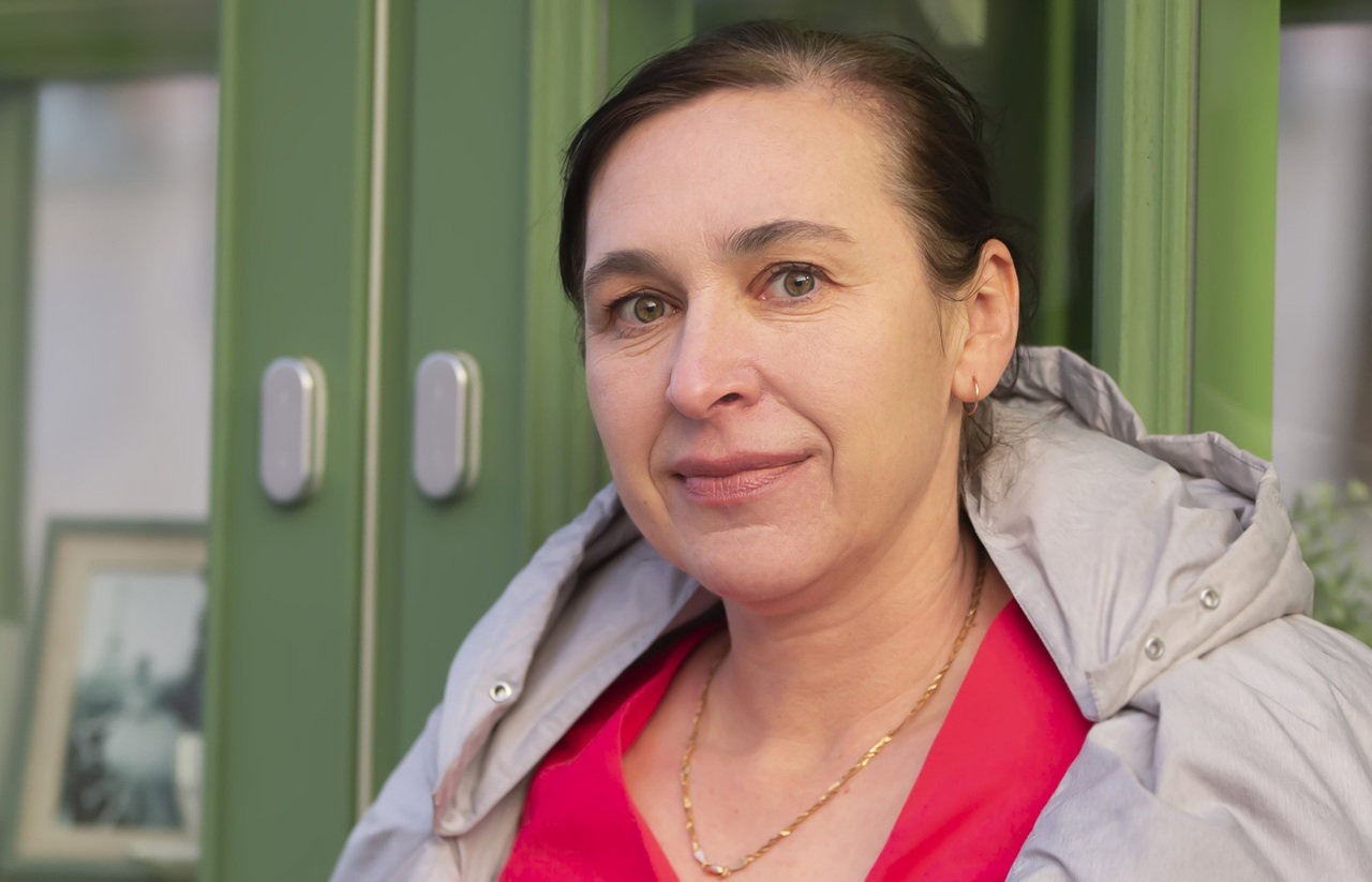 kobieta w wieku 45 plus w kurtce stoi na tle zielonej witryny z lekkim uśmiechem i smutkiem w oczach