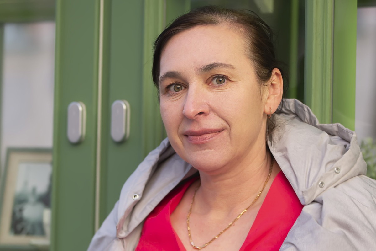 kobieta w wieku 45 plus w kurtce stoi na tle zielonej witryny z lekkim uśmiechem i smutkiem w oczach