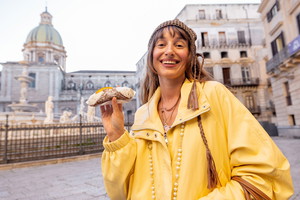 kobieta w wieku 30 plus w żółtej kurtce stoi na tle centrum Palermo z cannolo w dłoni