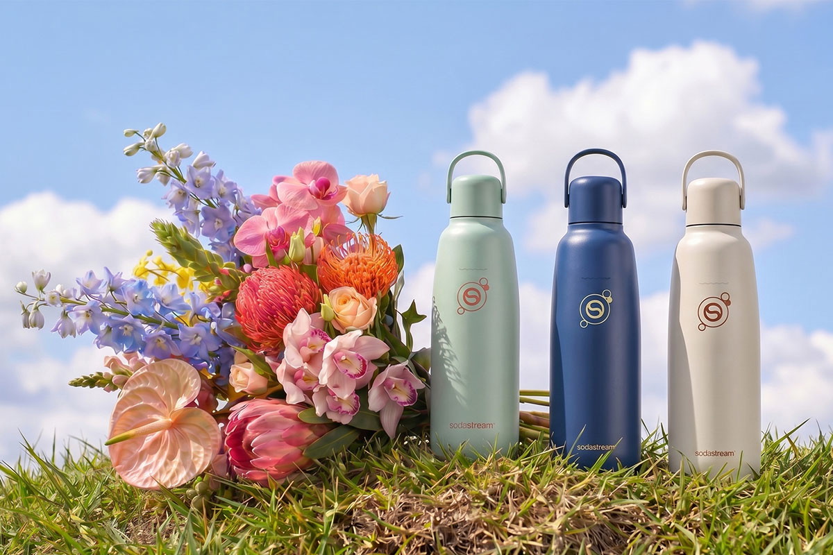 Trzy kolorowe butelki Fizz&Go Sodastream stoją obok bukietu kwiatów na trawie, pod błękitnym niebem - tytułowa, źrodło zdjęcia - AI
