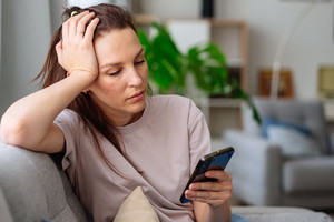 kobieta w wieku 30 plus siedzi na kanapie i przegląda Internet w komórce załamana
