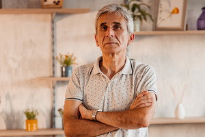mężczyzna w wieku 62 lat stoi z założonymi rękami na tle pokoju z poważną miną
