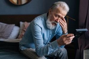 mężczyzna w wieku 65 plus siedzi na kanapie ze zdjęciem w ramce w ręku i ociera łzę