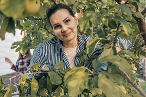 kobieta w wieku 45 plus ciemne włosy w koszuli w wąskie paski stoi wśród jabłoni z łagodną miną