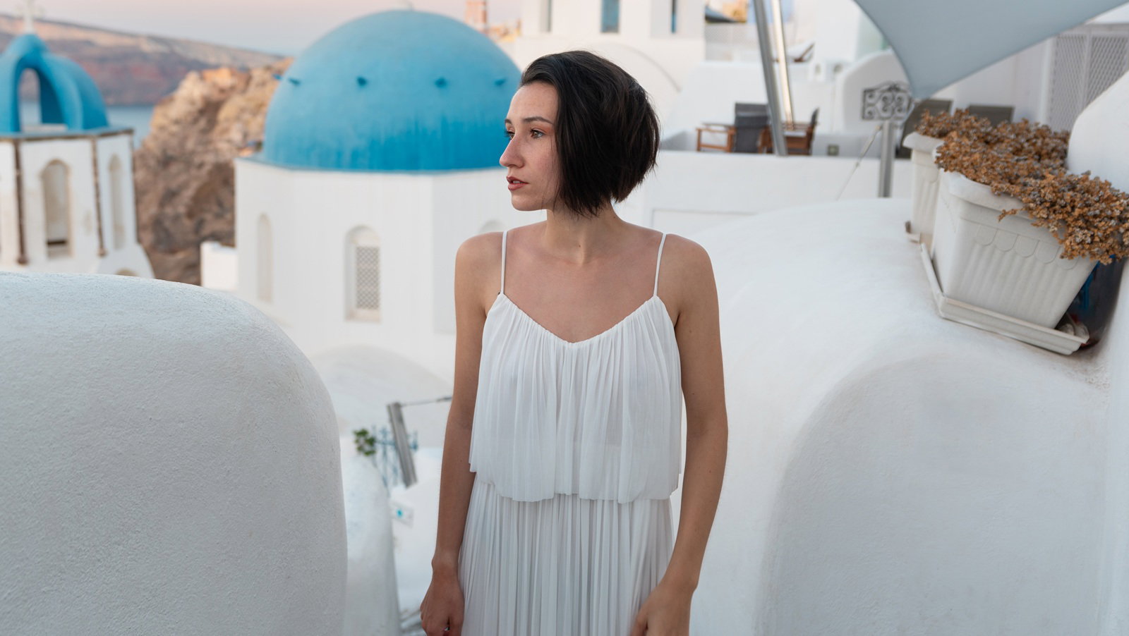 kobieta w wieku 30 plus w białej sukience stoi na tle pejzażu Santorini w Grecji smutna i zamyślona