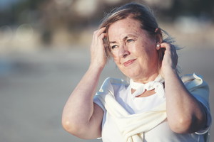 kobieta w wieku 55 plus stoi na plaży w bieli zamyślona i smutna poprawia włosy rozwiane przez wiatr