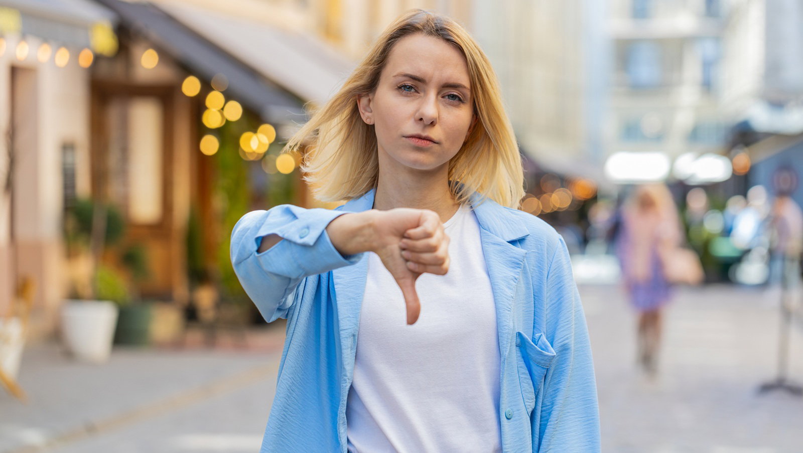 młoda kobieta w wieku 30 plus blondynka w niebieskiej marynarce stoi na ulicy niezadowlona i pokazuje kciuk w dół