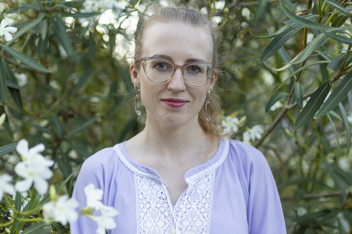 kobieta w wieku 30 plus w okularach i liliowej sukience stoi w ogrodzie z niepewnym uśmiechem