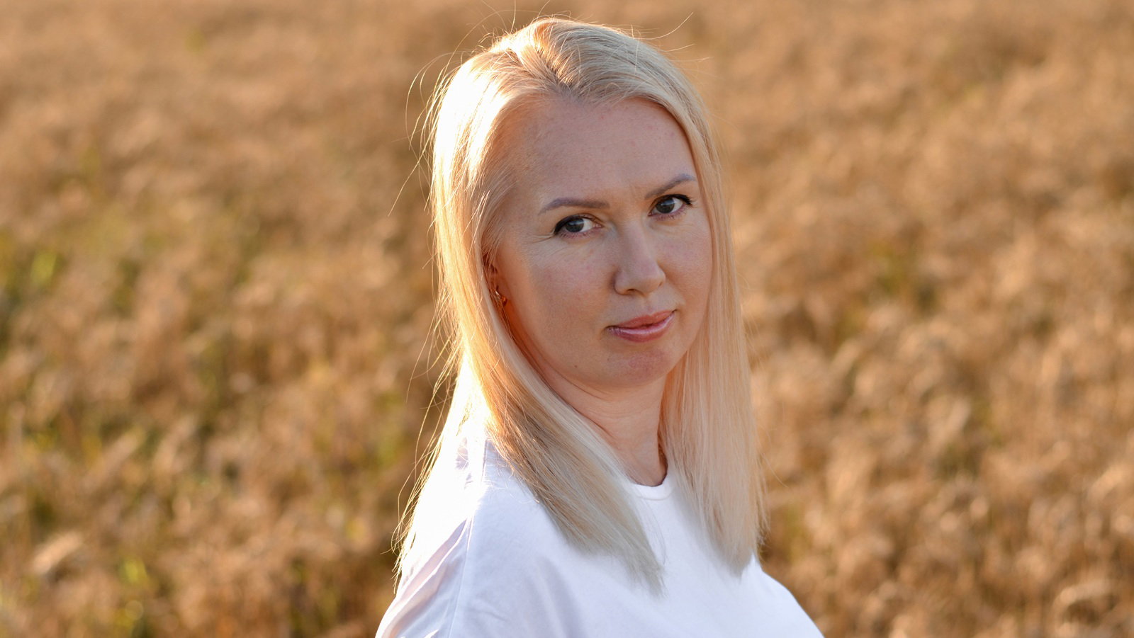 kobieta w wieku 35 plus blondynka w białej bluzce na tle pola stoi ze smutnym uśmiechem