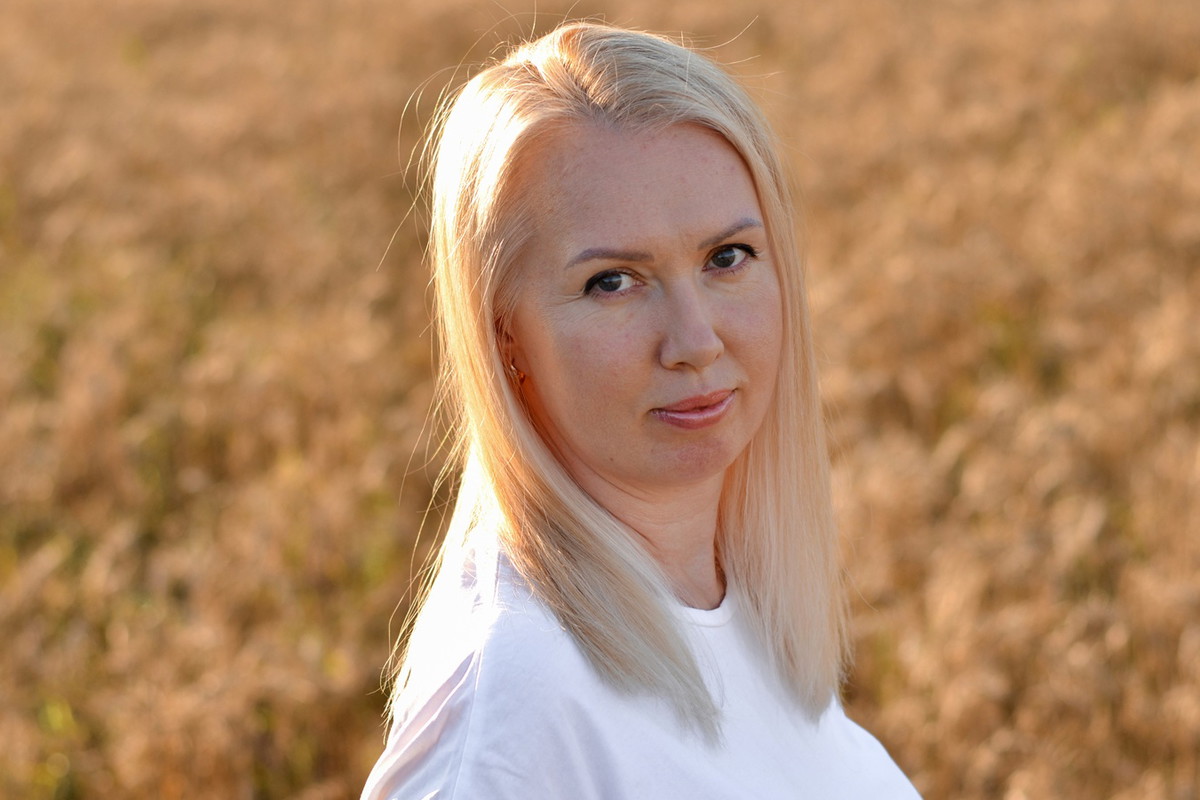 kobieta w wieku 35 plus blondynka w białej bluzce na tle pola stoi ze smutnym uśmiechem