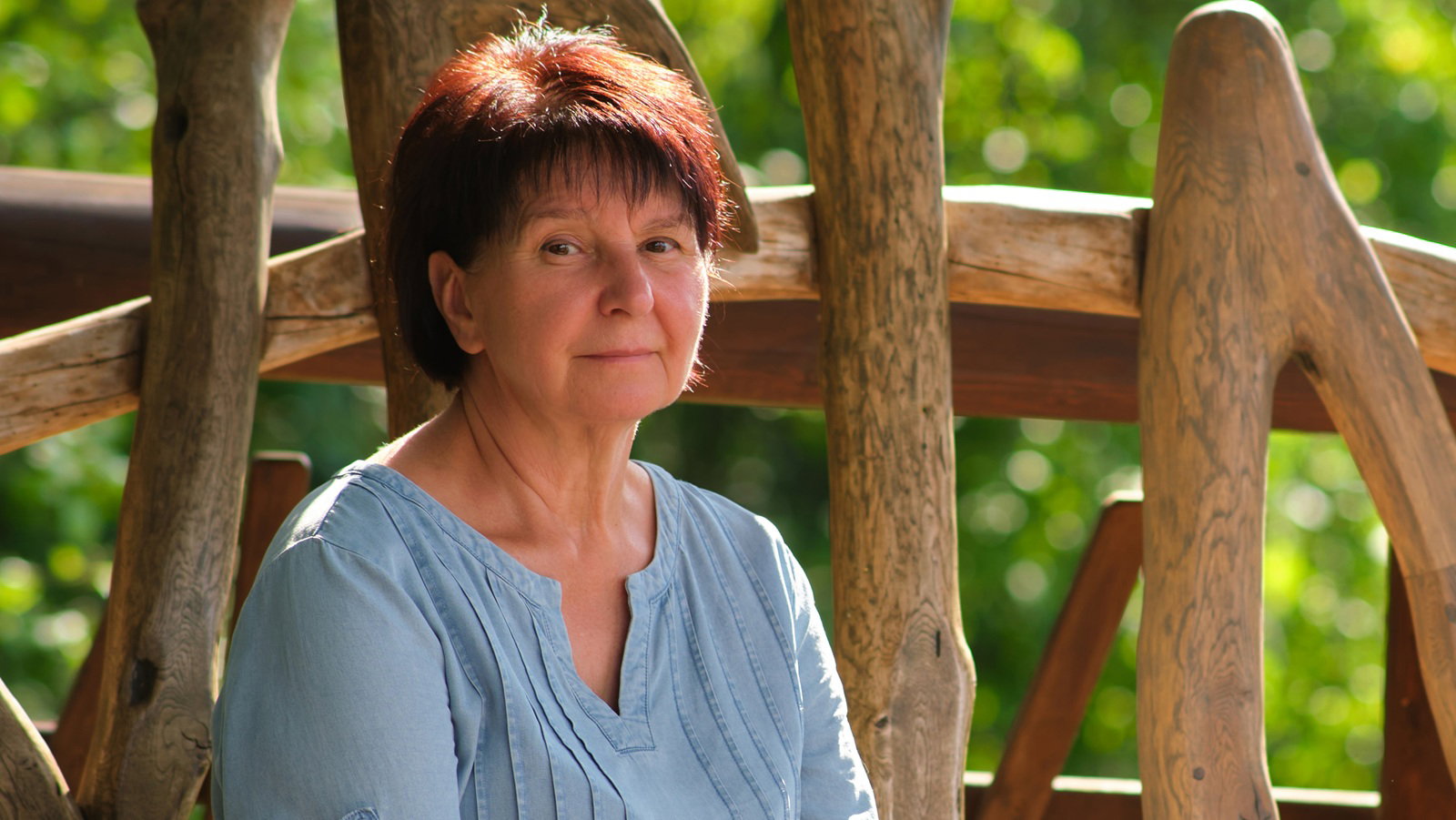 kobieta w wieku 60 plus siedzi przy drewnianym płocie w niebieskiej bluzeczce zamyślona i smutna