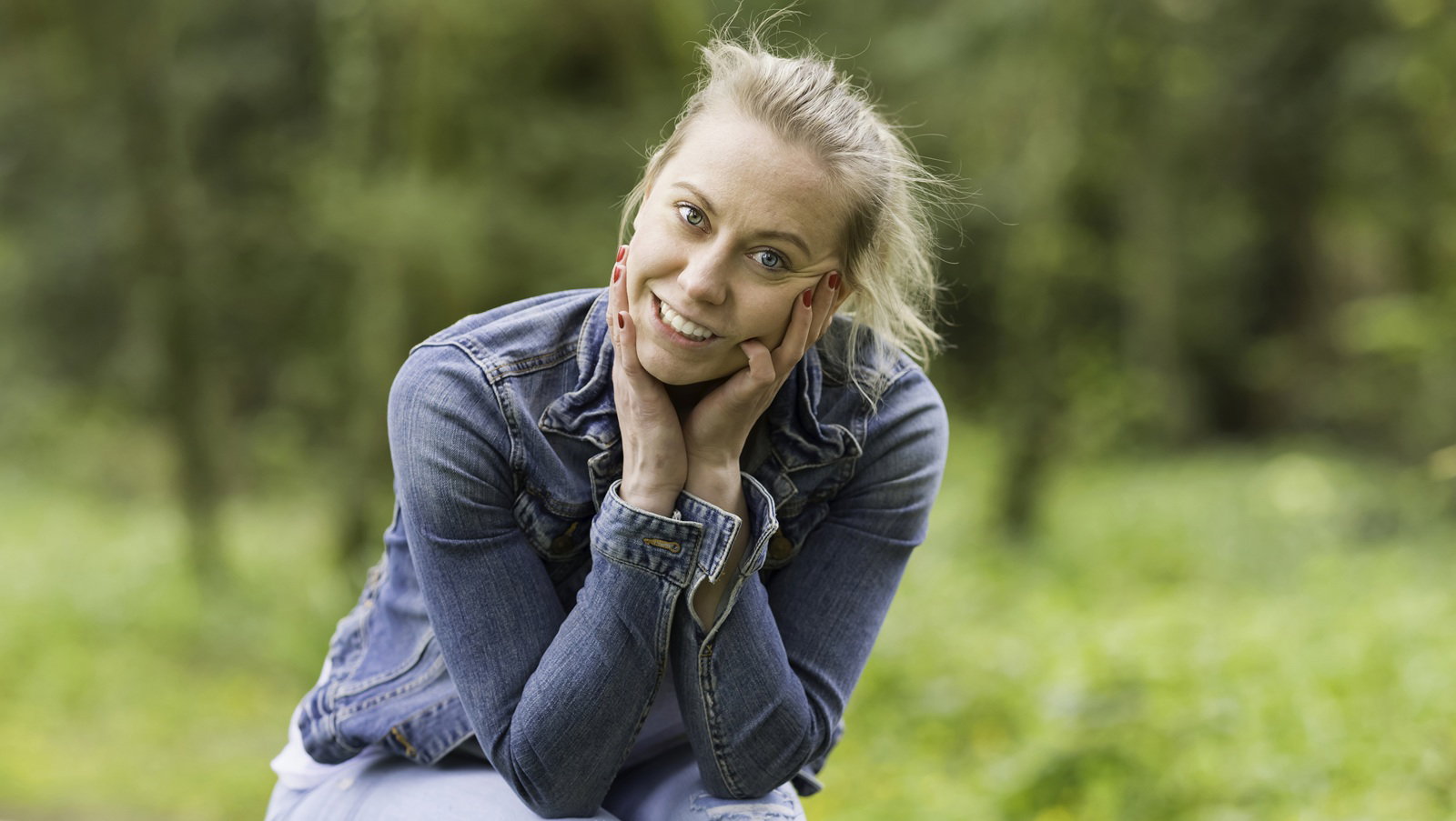 kobieta w wieku 30 plus blondynka siedzi na pieńku na tle lasu w jeansowej kurtce zadowolona i rozbawiona