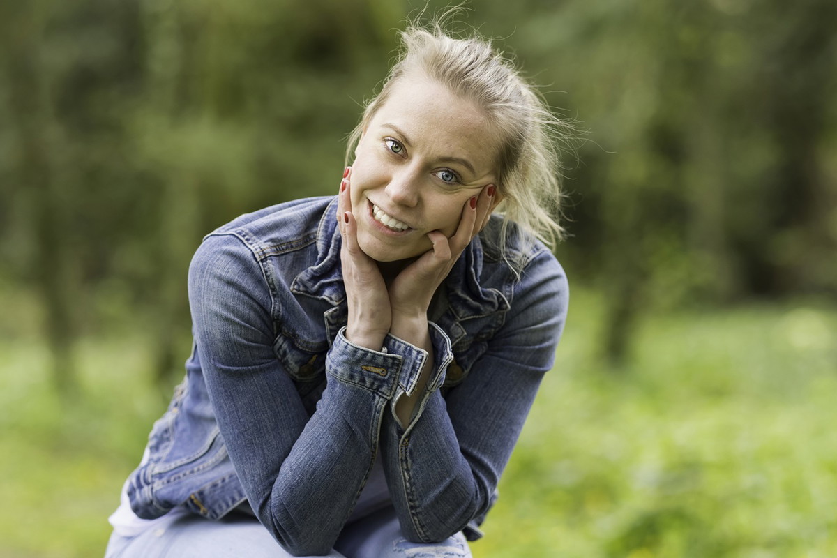 kobieta w wieku 30 plus blondynka siedzi na pieńku na tle lasu w jeansowej kurtce zadowolona i rozbawiona