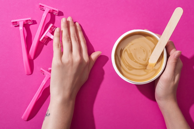cropped view of woman moving away disposable razors and taking depilation wax in cup with stick on pink background