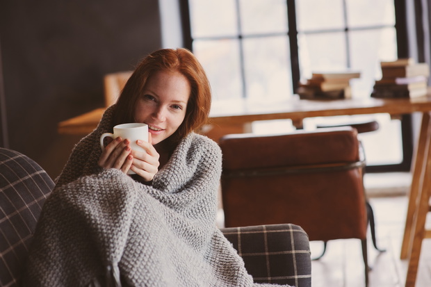 young sick woman healing with hot drink at home on cozy couch, wrapped in knitted blanket
