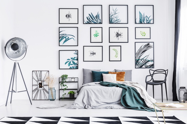 Real photo of a wide bedroom standing between a chair and a shelf, lamp and a clock in a spacious bedroom with triangle rug and wall full of paintings