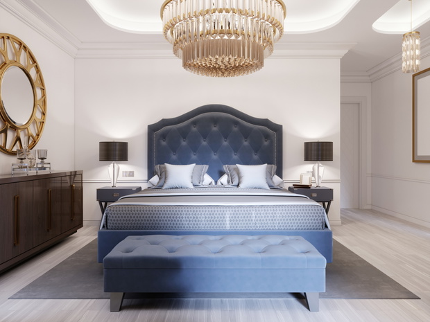 Modern bed in classic blue style with bedside table and lamp. Large glass chandelier over. A dresser with a decor and a golden mirror above. Modern bedroom.