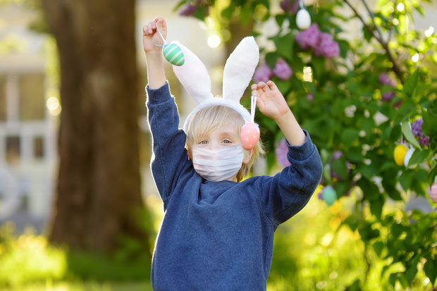 Pretty little boy wearing bunny ears and disposable medical face mask hunting for easter eggs in spring park on Easter day during coronavirus outbreak.