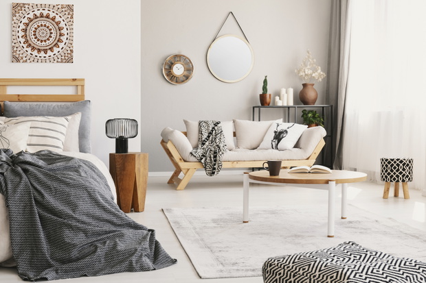 Small flat interior with bed, window with curtains, coffee table with mug and book placed on carpet, lounge with blanket and cushion and clock and mirror on the wall in real photo