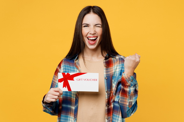 Young woman wear blue shirt beige t-shirt hold store gift certificate coupon voucher card do winner gesture wink blink eye isolated on plain yellow background studio portrait People lifestyle concept