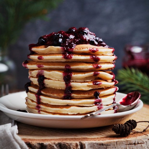 Stack of american pancakes or fritters with strawberry and blueberry jam in white plate on wooden rustic table decorated Christmas tree, delicious dessert for breakfast in winter, vintage style