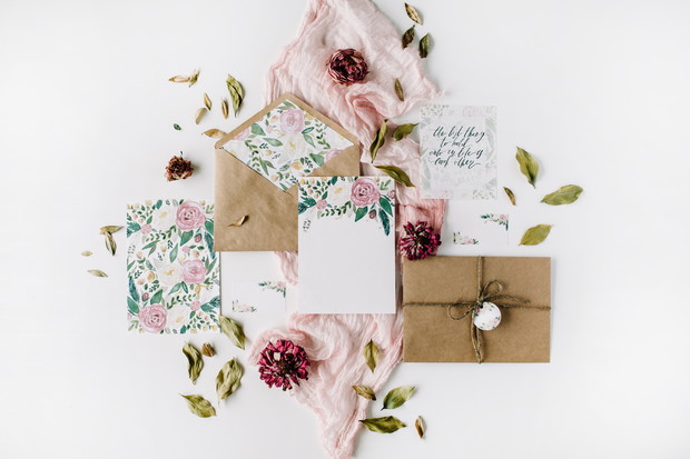 Workspace. Wedding invitation cards, craft envelopes, pink and red roses and green leaves on white background. Overhead view. Flat lay, top view