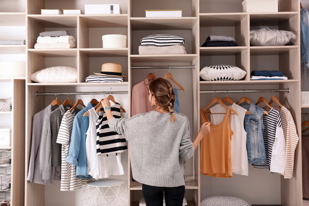 Woman choosing outfit from large wardrobe closet with stylish clothes and home stuff