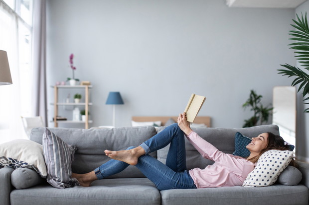 Young beautiful woman is reading a book while lying on a sofa in the living room.
