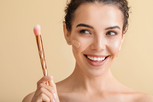 Beautiful young woman with foundation on her face against color