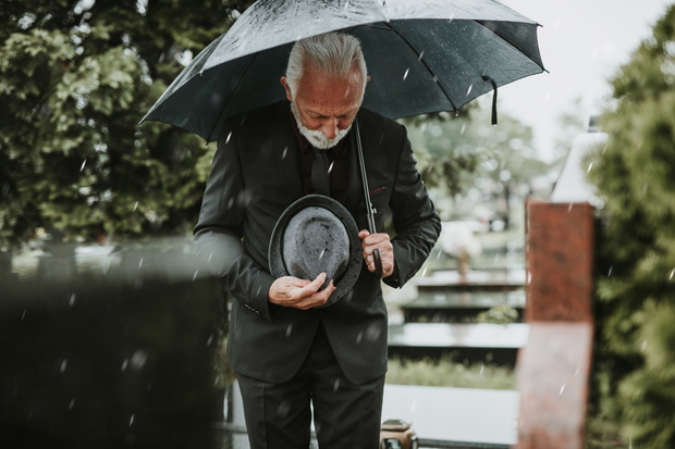 Elegant sad elderly man standing on the rain with umbrella and g