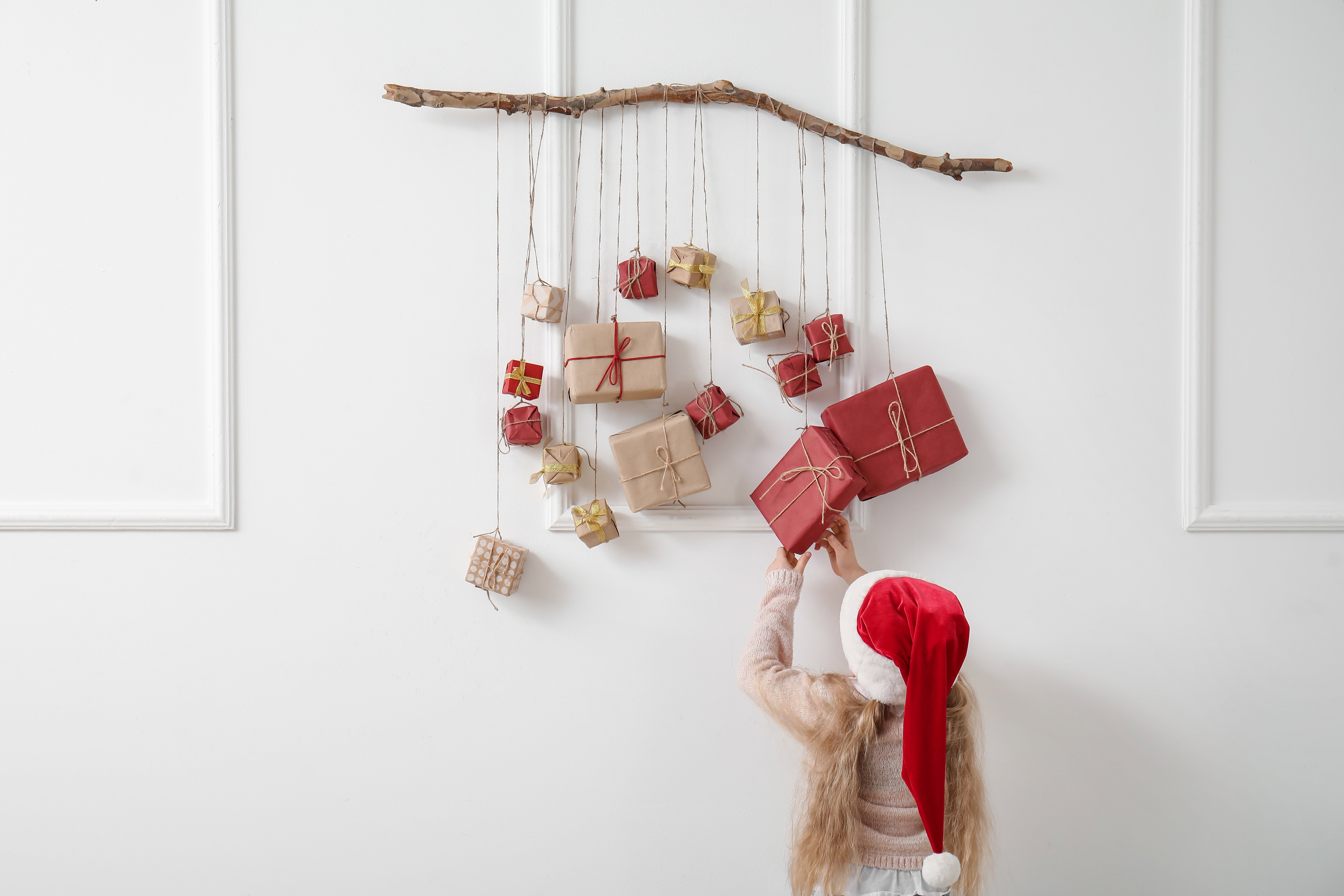 Cute little girl in Santa hat and Christmas advent calendar hanging on light wall