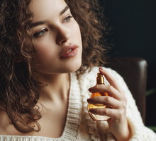 Close up portrait of young beautiful sensual curly woman using, holding luxury perfume in orange glass bottle. Model wearing warm winter knitted sweater. Copy, empty space for text