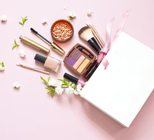 Various cosmetic products eye shadow powder lip gloss blush mascara brush for makeup and white spring flowers in a white gift bag on a pink pastel background top view with copy space