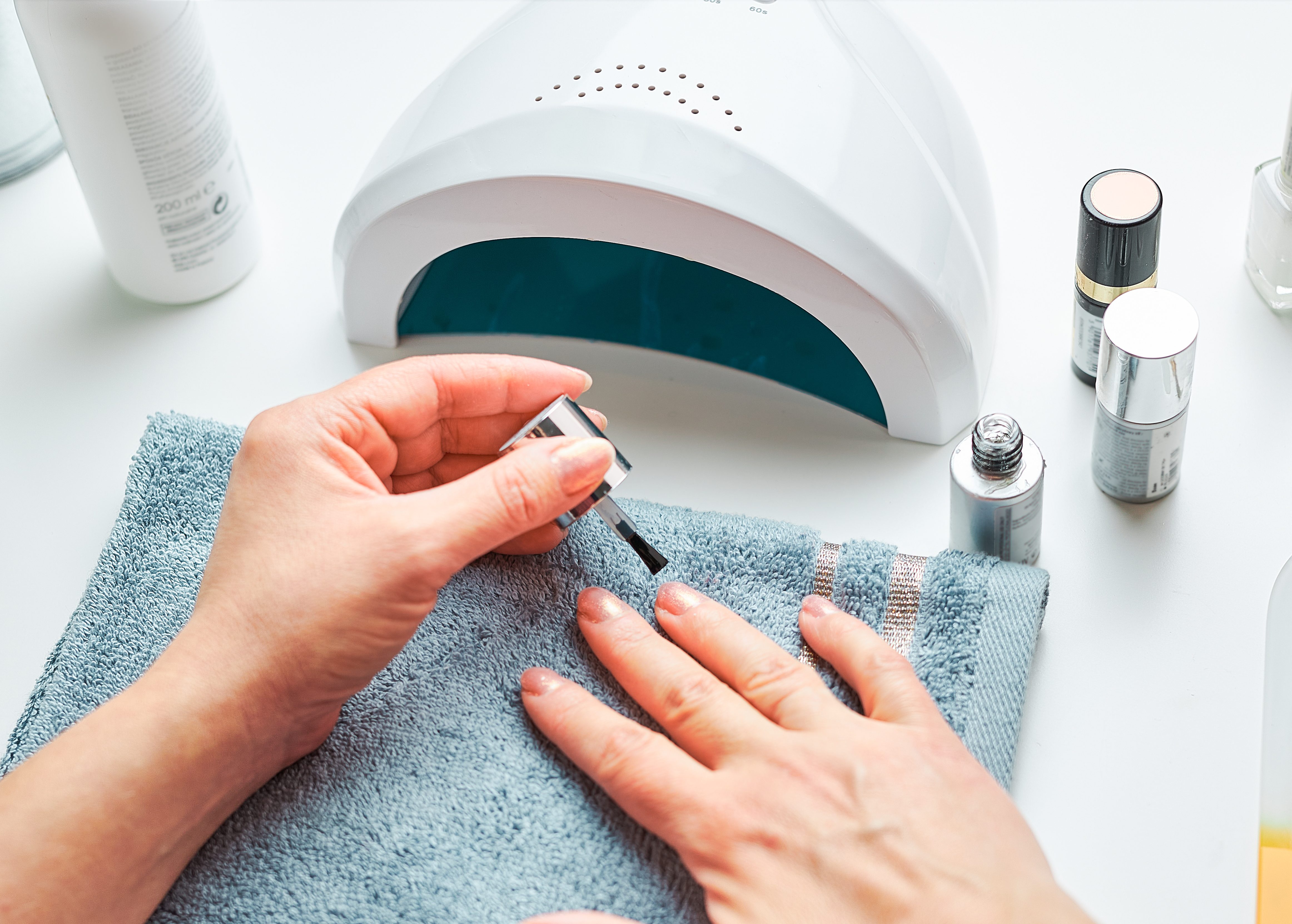 Woman applying gel hybrid polish using UV lamp. Beauty wellness spa treatment concept. Cosmetic products, UV lamp, green leaves on white table. Spa, manicure, skin care concept. Flat lay, overhead view