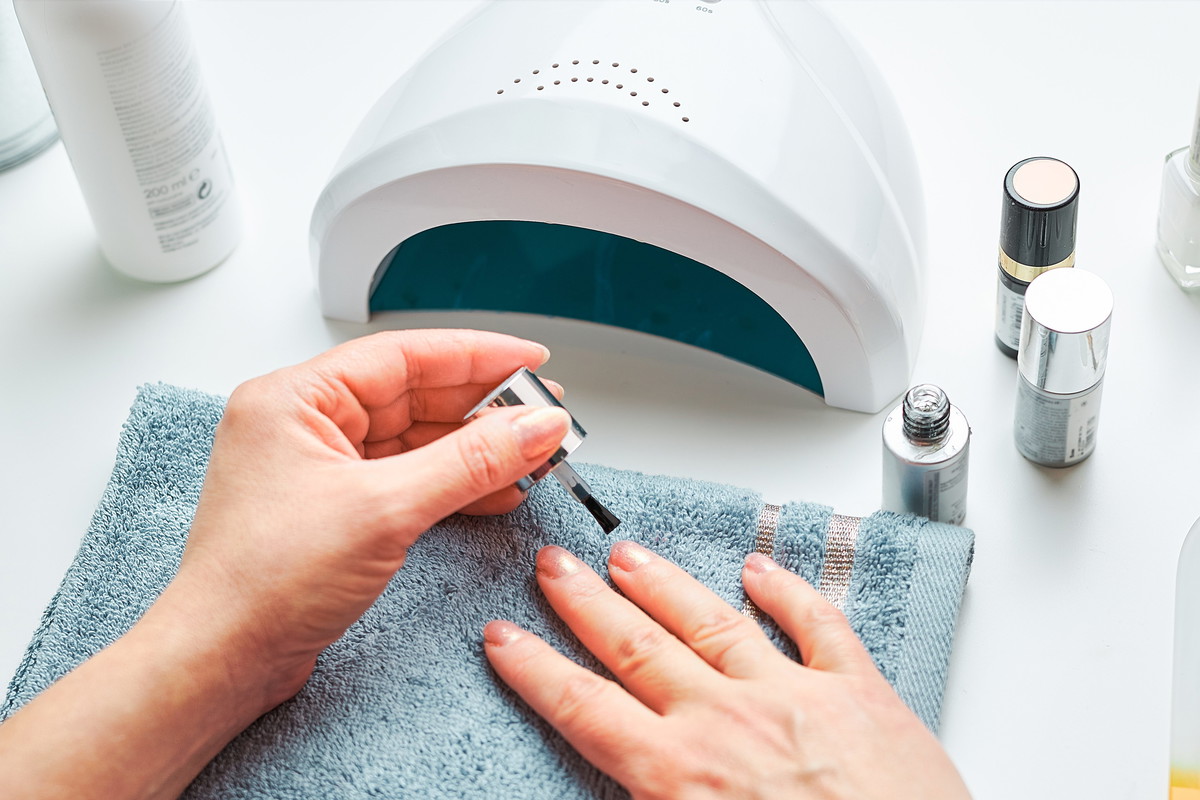 Woman applying gel hybrid polish using UV lamp. Beauty wellness spa treatment concept. Cosmetic products, UV lamp, green leaves on white table. Spa, manicure, skin care concept. Flat lay, overhead view