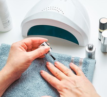 Woman applying gel hybrid polish using UV lamp. Beauty wellness spa treatment concept. Cosmetic products, UV lamp, green leaves on white table. Spa, manicure, skin care concept. Flat lay, overhead view