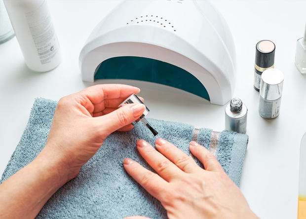 Woman applying gel hybrid polish using UV lamp. Beauty wellness spa treatment concept. Cosmetic products, UV lamp, green leaves on white table. Spa, manicure, skin care concept. Flat lay, overhead view