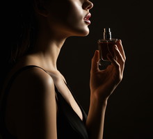 Woman in a black dress holding a perfume bottle close to her neck, captured with dramatic low light for a sensual and intimate mood.