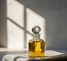 A glass bottle with golden liquid sits atop a folded gray towel, bathed in sunlight casting shadows on a white surface.