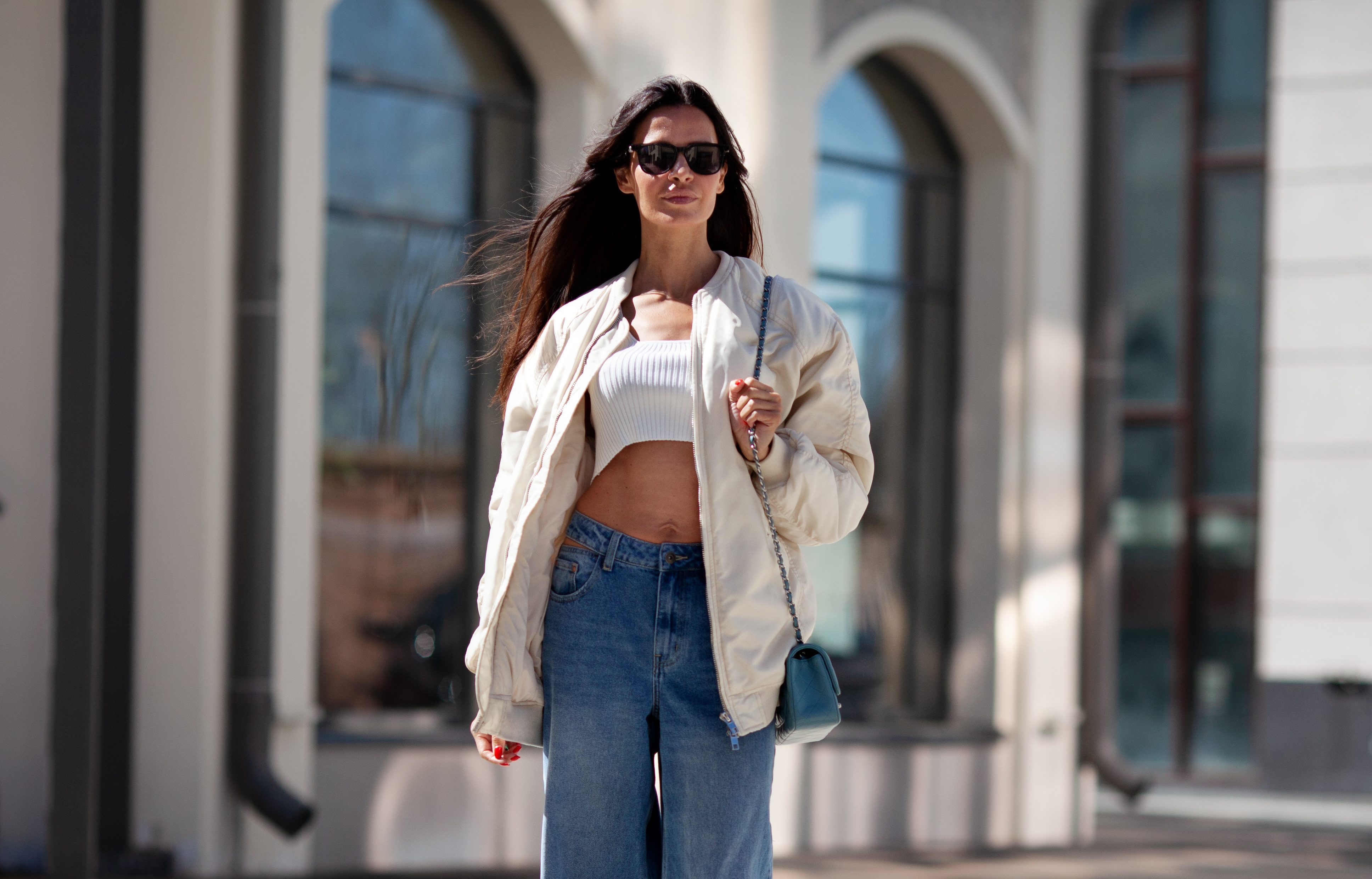 Fashionable woman with long flowing hair wearing trendy white bomber jacket, short Tshirt, wide jeans, posing on street of city. Trends clothing of spring and summer