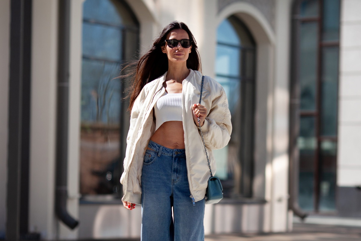 Fashionable woman with long flowing hair wearing trendy white bomber jacket, short Tshirt, wide jeans, posing on street of city. Trends clothing of spring and summer