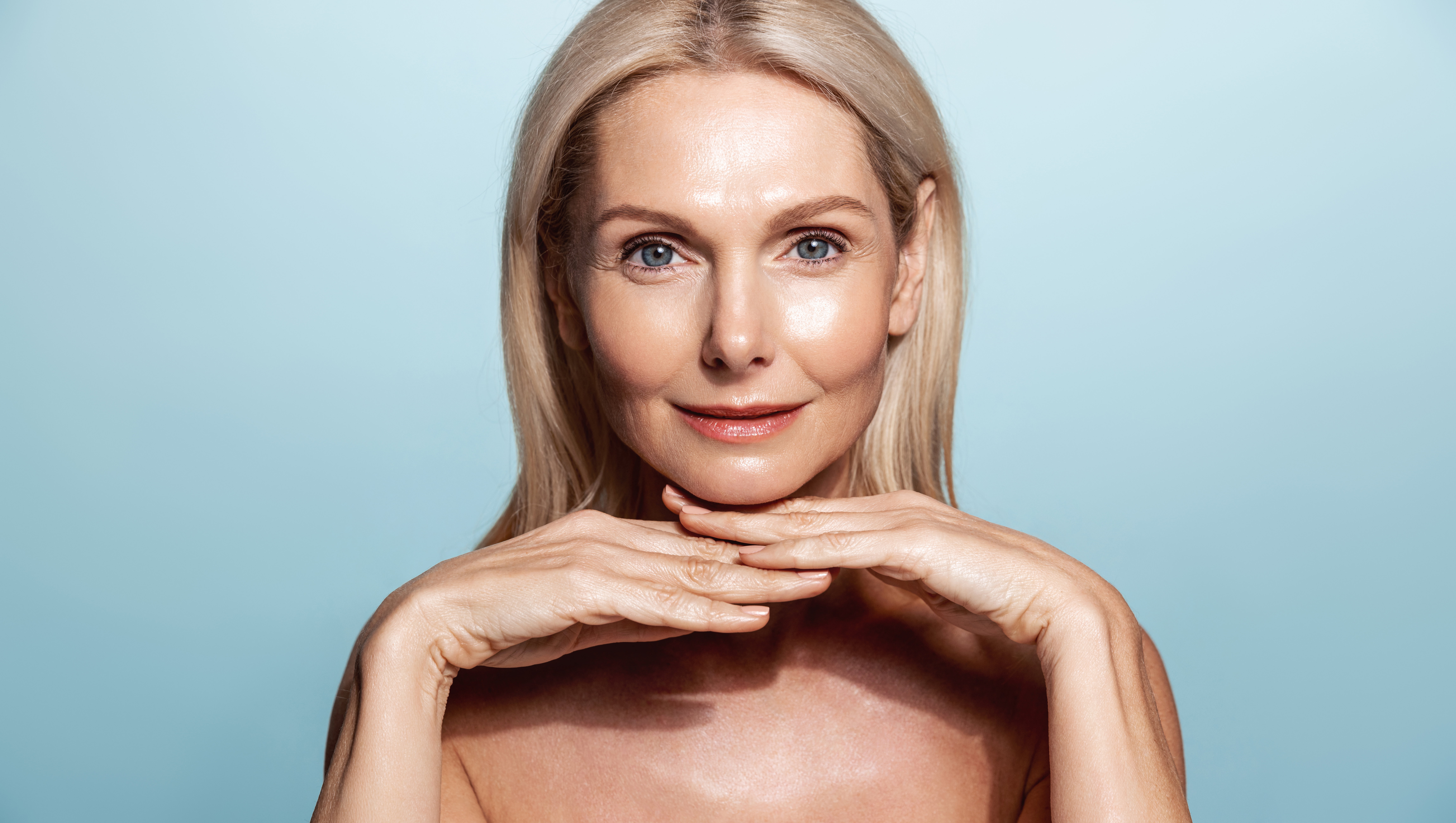 Beautiful middle aged woman with perfect face glow, using spa, anti-aging lifting cream and nourishment cosmetic products, blue background
