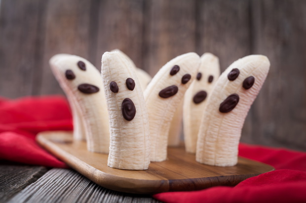 Homemade halloween scary banana ghosts monsters with chocolate faces. Healthy natural vegetarian snack funny dessert recipe for party decoration on vintage wooden table background and red fabric