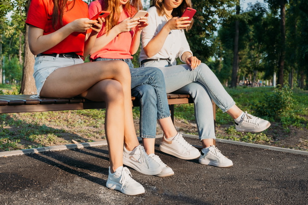 Diverse three girl friends using their phones outdoors. Group gen z young people using mobile smartphone sitting on bench in summer park