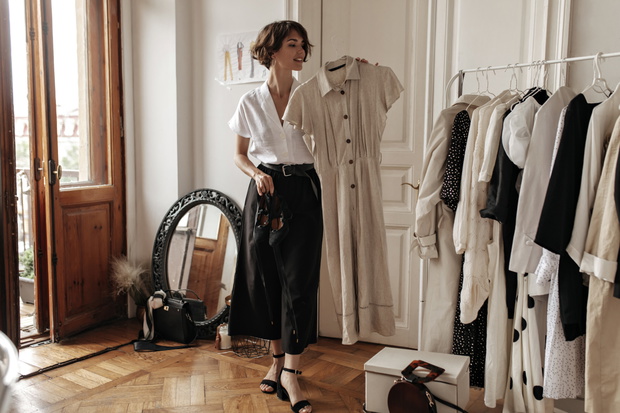 Cheerful short-haired brunette woman in trendy midi skirt, white blouse poses in cozy dressing room, holds beige linen dress and shoes