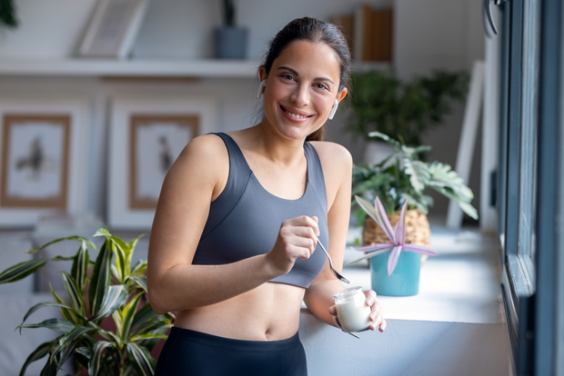 Beautiful sporty woman eating yogurt while listening music with earphone standing near the window at home.