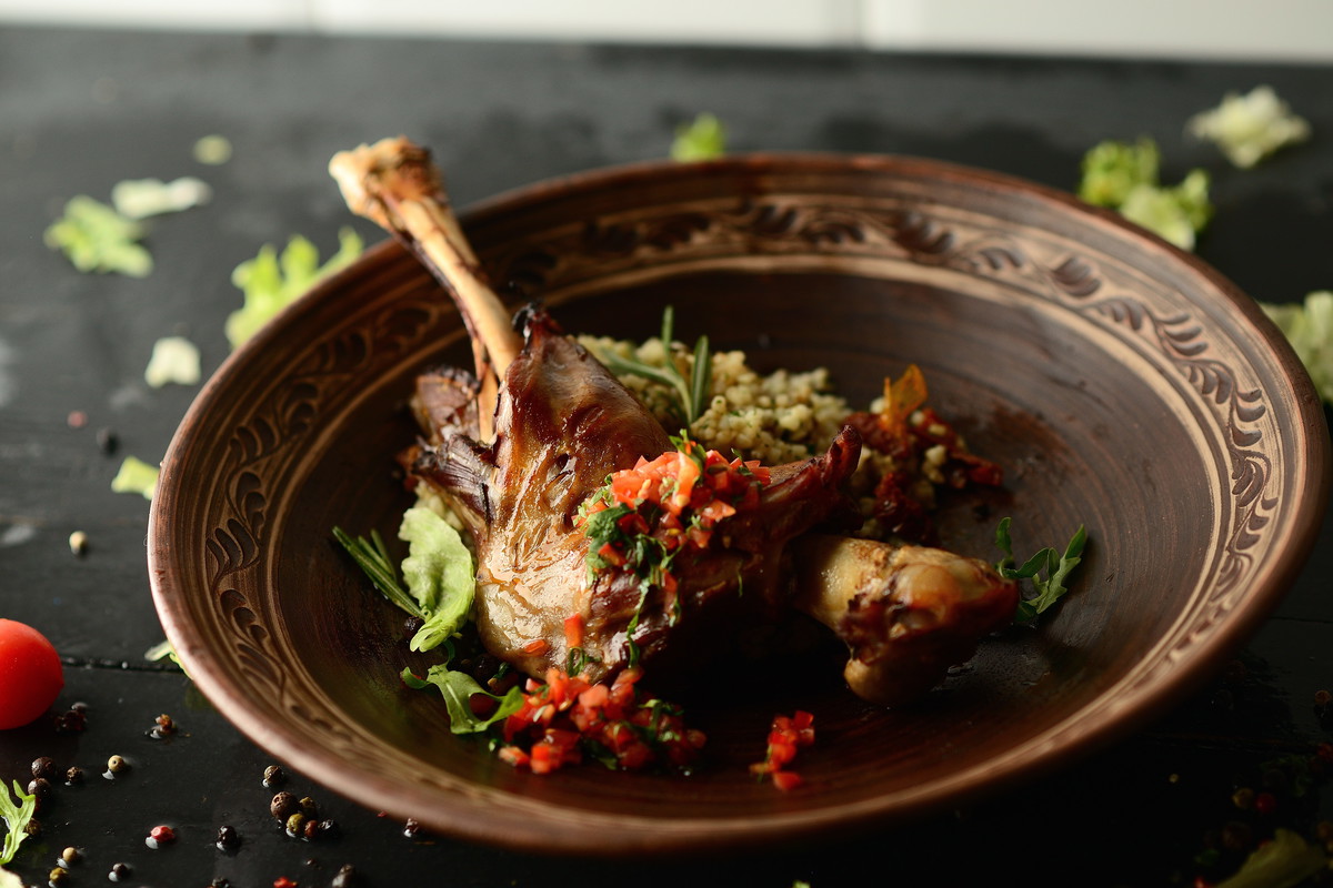 Roasted goose leg, sauce, herbs and tomatoes in a clay bowl on wooden background
